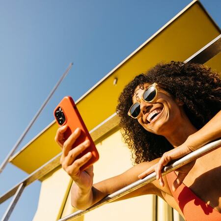 Woman smiling at her phone while leaning on a balcony railing near the beach on a sunny day