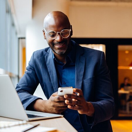 Man in business attire texting on his phone while working at a cafe with an open laptop and notebook