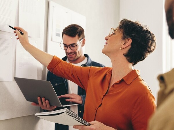 Person pointing to documents on a wall while holding a notebook, collaborating with another person using a laptop in an office setting
